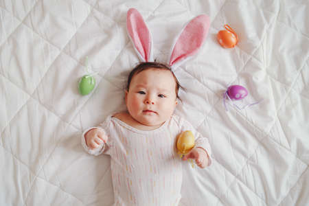 Cute adorable mixed Asian baby wearing pink Easter bunny ears lying on bed in bedroom with colorful...