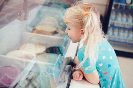 Caucasian funny blonde child girl looking at ice cream in shop window trying to choose one. Kid with puzzled emotional face expression. Candid childhood lifestyle. Tasty frozen summer food.の写真素材