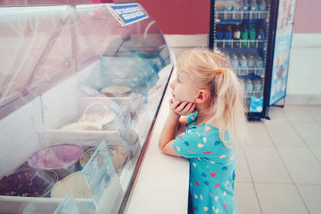 Caucasian funny blonde child girl looking at ice cream in shop window trying to choose one. Kid with puzzled emotional face expression. Candid childhood lifestyle. Tasty frozen summer food.の写真素材