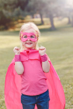 Cute adorable preschool Caucasian child playing superhero in costume. Girl kid wearing pink mask and cape having fun outdoors in park. Happy active childhood. Feminism and power concept.の写真素材