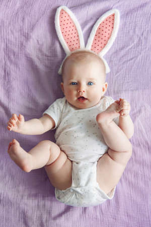 Cute adorable white Caucasian baby girl wearing pink Easter bunny ears lying on pink purple bed in...