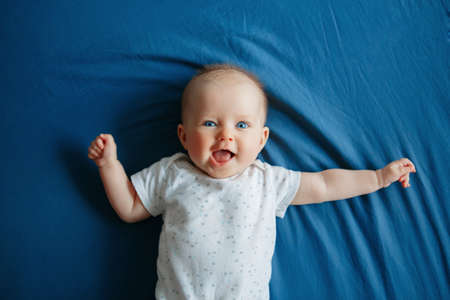 Portrait of cute adorable smiling white Caucasian baby girl boy with blue eyes four months old lying on bed in bedroom looking at camera. View from top above. Happy childhood lifestyle.の写真素材
