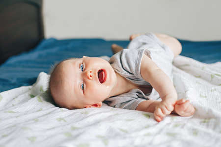 Portrait of crying screaming drooling white Caucasian baby girl boy four months old lying on bed in bedroom on tummy. Natural emotion expression. Childhood lifestyleの写真素材