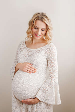 Portrait of smiling white Caucasian blonde pregnant woman in white lacy dress touching her belly. Expecting beautiful young middle age female lady. Healthy happy parenthood lifestyle concept.の写真素材