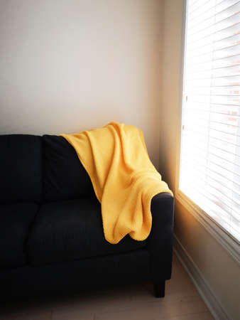 Bright yellow knitted blanket lying on dark black brown sofa couch near window with natural day light through blindsの写真素材