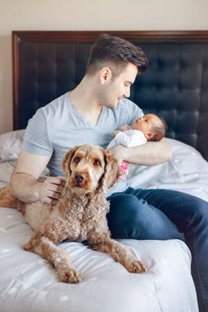 Portrait of young Caucasian father with mixed race Asian Chinese newborn baby. Dog pet lying on bed. Man parent holding child daughter son. Authentic lifestyle moment. Single dad family life. の写真素材