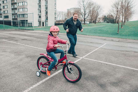Smiling excited Caucasian father training helping girl daughter to ride bicycle. Preschooler child kid in pink helmet with bike on backyard road outside on spring day. Seasonal child outdoor activityの写真素材