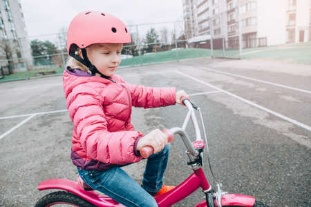 Portrait of smiling Caucasian preschooler girl riding pink bike bicycle in helmet on backyard road outside on spring day. Seasonal child activity concept. Healthy childhood lifestyleの写真素材