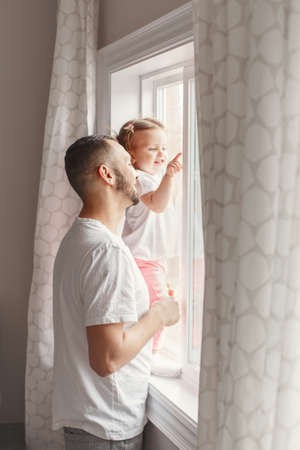 Portrait of Caucasian young father dad with toddler girl daughter looking into window waiting expecting someone. Child staring watching from home outside into street and pointing with her finger.の写真素材