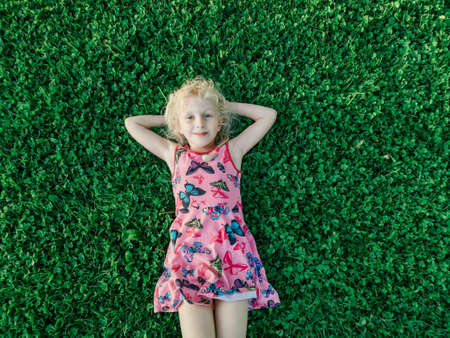 Portrait of cute funny adorable blonde Caucasian smiling girl in red pink dress lying on grass enjoying spring summer sunny day. Happy childhood lifestyle concept. View from top above.の写真素材