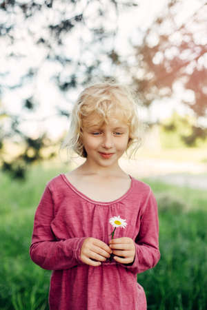 Portrait of cute beautiful blonde Caucasian girl in red pink dress holding white daisy flower in her hands. Happy adorable child kid with messy untidy hair enjoying summertime. Lifestyle childhood.の写真素材