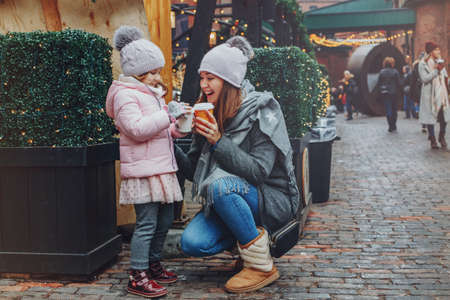 Mother with child girl drinking hot chocolate coffee at Christmas market celebrating New Year holiday. Family outdoor winter activity. Mom and daughter spend time together. Authentic lifestyle.の写真素材