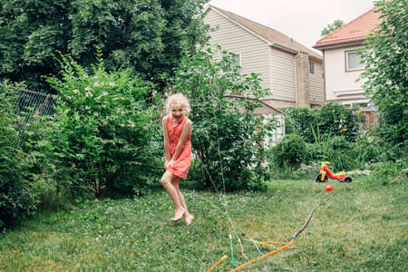 Preschool kids girl splashing with gardening hose sprinkler on backyard during summer day. Child playing with water outside at home yard. Candid authentic real life moment of funny family activity.の写真素材