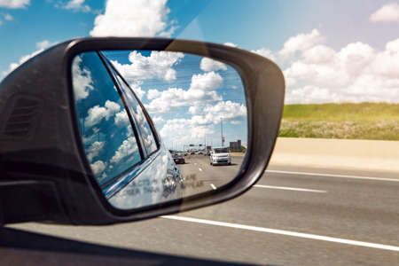 Closeup of rear back car mirror with beautiful landscape midday view of  Toronto city highway street road. Cars traffic during sunny day with white clouds in blue sky reflected in vehicle glass.の写真素材
