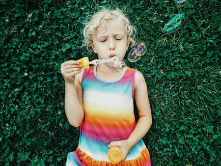 Candid portrait of cute funny little blonde Caucasian child girl lying on ground grass blowing soap...