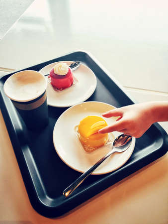 Closeup of kids child hand trying to steal tasty cakes from plate on tray. Impatient person taking food dessert on dish. Tasty delicious pastry with coffee cup in cafeteria cafe.の写真素材
