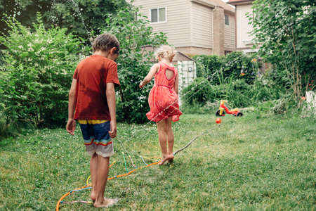 Kids friends splashing with gardening hose sprinkler on backyard on summer day. Children jumping  playing with water outside. Candid authentic real life moment of funny activity. View from back.の写真素材
