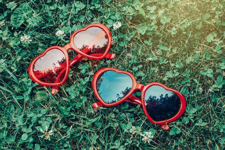 Two colorful funny silly red heart shaped sunglasses lying in green grass on summer day. View from top above. Toned with retro vintage hipster filters.の写真素材