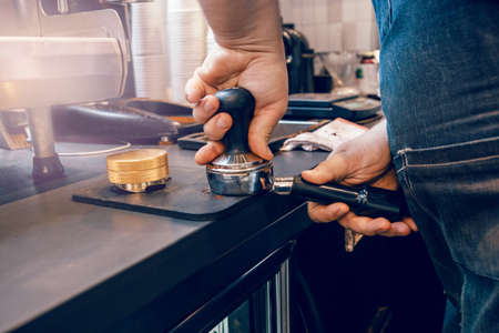 Closeup of barista hands using tamper to press ground coffee into portafilter to make espresso hot...