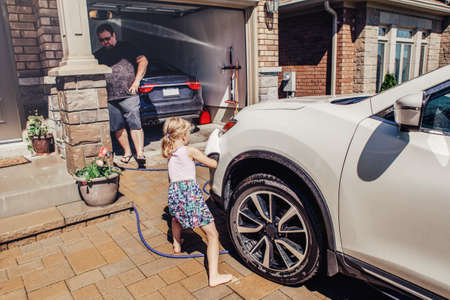 Cute preschool little Caucasian girl helping father wash car on driveway in front house on summer day. Child wiping cleaning machine bumper. Kids home errands duty chores responsibility concept.の写真素材