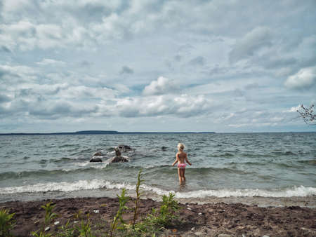 Cute Caucasian preschool blonde girl in pink swimsuit walking in Georgian Bay Canadian Ontario lake water. Concept of harmony with nature, happy healthy active childhood. View from back.の写真素材
