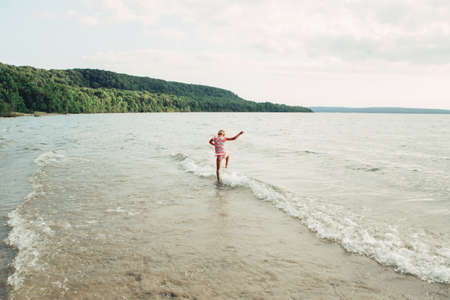 Cute funny Caucasian girl walking in lake river waves at sunset. Child jumping water on beach. Authentic real lifestyle happy childhood. Summer fun outdoor aquatic activity. View from back.の写真素材