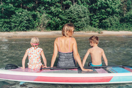 Caucasian woman parent sitting on paddle sup surfboard in water talking to kids children. Modern outdoor family activity. Individual aquatic recreation sport hobby. View from back.の写真素材
