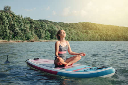Middle age Caucasian woman practicing yoga on paddle sup surfboard at sunset. Female stretching doing workout on lake water. Modern individual hipster outdoor summer sport activity.の写真素材