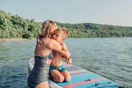Caucasian woman mother hugging comforting crying sad son boy child on paddle sup surfboard in water. Mom Modern outdoor family activity sport hobby.の写真素材