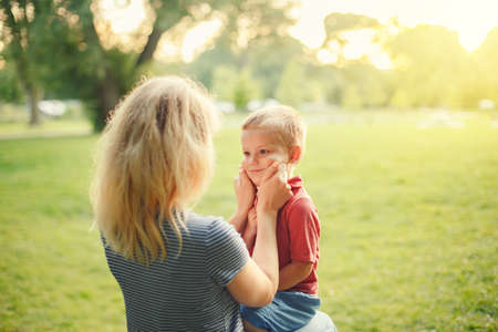 Young Caucasian mother pulling boy toddler cheeks. Mom sitting together with son on summer day in park and showing her love and affection. Happy family childhood lifestyle. Cute authentic moment.の写真素材