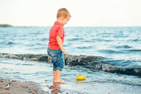 Happy Caucasian red-haired toddler child boy putting yellow paper boat in water on lake sea ocean shore at evening sunset or morning. Kid playing on beach. Happy lifestyle childhood concept.の写真素材