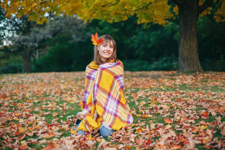 Beautiful Caucasian woman sitting on ground in autumn fall park with red yellow maple leaf in hair. Girl wrapped in woollen warm blanket. Cozy autumnal seasonal mood feeling concept.の写真素材