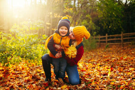 Caucasian young mother hugging playing with cute adorable toddler boy son in autumn fall park outdoor with yellow orange leaves trees. Thanksgiving autumnal seasonal concept. の写真素材