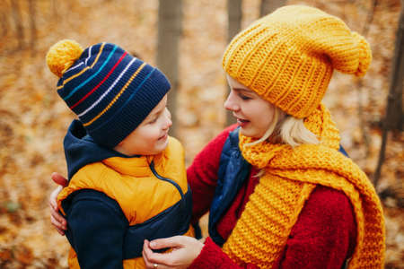 Caucasian young mother hugging talking to cute adorable toddler boy son in autumn fall park outdoor with yellow orange red tree leaves. Happy family. Autumnal seasonal activity outdoor.の写真素材