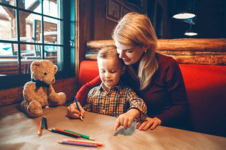 Portrait of young Caucasian mother and son sitting in restaurant drawing with colored pencils on craft brown paper. Family together in cafe. Lifestyle authentic real people. Toned with filters.の写真素材