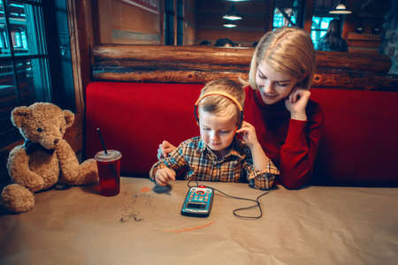 Portrait of young blonde Caucasian mother with toddler boy son sitting in cafe restaurant and playing electronic digital gadget device toyの写真素材