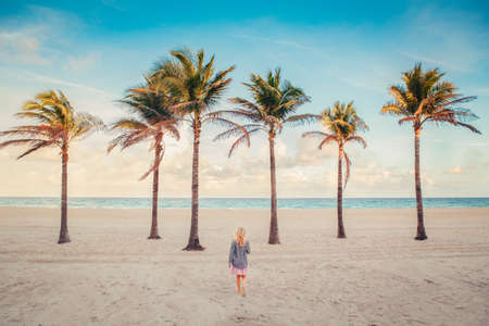 Little blonde Caucasian girl walking on empty Hollywood ocean beach in Florida. Child among tall palm trees on summer sunny day at sunset. View from back. Travel vacation kids holiday concept.の写真素材
