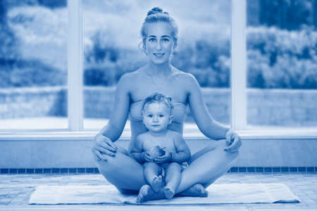 Caucasian mother and baby daughter doing fitness exercises yoga sitting in lotus pose looking at camera. Toddler eating apple. Sport family together. Toned with classic blue 2020 color.の写真素材