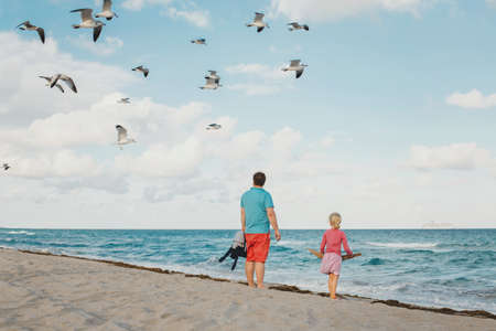 Father and daughter standing on sea ocean beach and watching seagulls in sky above. Dad and child girl travelling spending time together at summer day on shore coast. View from back.の写真素材