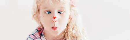 Cross-eyed kid looking at red heart sticker on nose. Funny hilarious white Caucasian cute adorable child girl with blue eyes. Valentine Day holiday concept. Web banner header for website.の写真素材