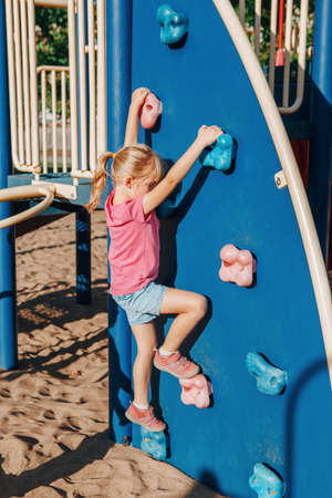 Little preschool girl climbing rock wall at playground outside on summer day. Happy childhood lifestyle concept. Seasonal outdoor activity for kids. Strong girl female power.の写真素材