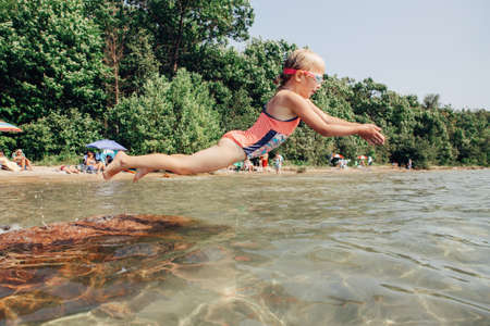 Cute funny Caucasian girl diving in lake with underwater goggles. Child swimming in water on Awenda beach. Authentic real lifestyle happy childhood. Summer fun outdoor seasonal activity.の写真素材