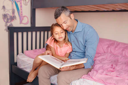 Dad reading book to pre-teen daughter girl. Happy family of two sitting on bed in bedroom. Smiling father and child at home spending time together. Education and authentic lifestyle childhood.の写真素材