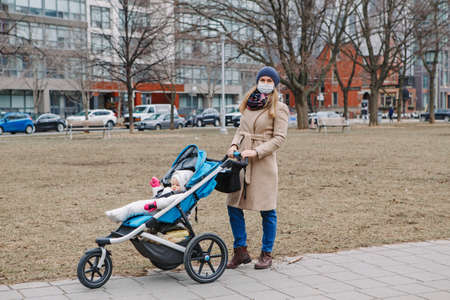 Young Caucasian mother in surgical mask walking with baby outdoor in Toronto. Protective face mask...