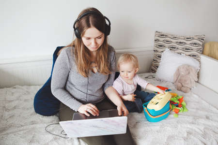 Young Caucasian mother blogger with baby working on laptop from home. Workplace of freelance woman student with kid toddler. Stay at home single mom earning money at online job.の写真素材