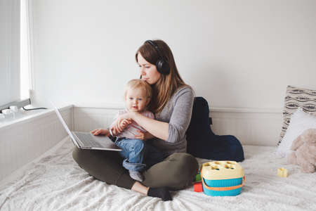 Young Caucasian mother blogger with baby working on laptop from home. Workplace of freelance woman student with kid toddler. Stay at home single mom earning money at online job.の写真素材