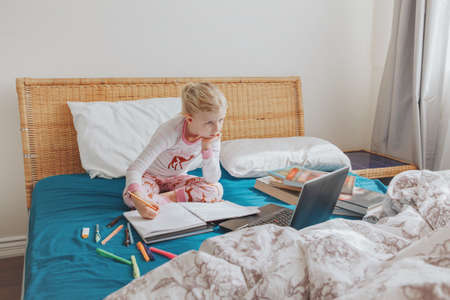Caucasian girl child sitting in bed and learning online on laptop Internet. Virtual class lesson on video during self isolation at home. Distant remote video education. Modern school study for kids.の写真素材