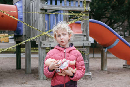 Sad upset Caucasian girl with baby toy on closed playground outdoor. Kids play area locked with...
