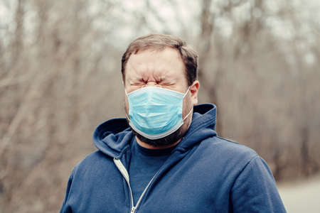 Caucasian young middle age man in sanitary face mask sneezing coughing outdoor. Person protecting from dangerous spread of virus. Coronavirus COVID-19 disease quarantine.の写真素材