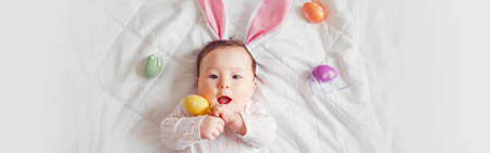 Cute adorable Asian baby wearing pink Easter bunny ears. Infant kid lying on bed with colored Easter...